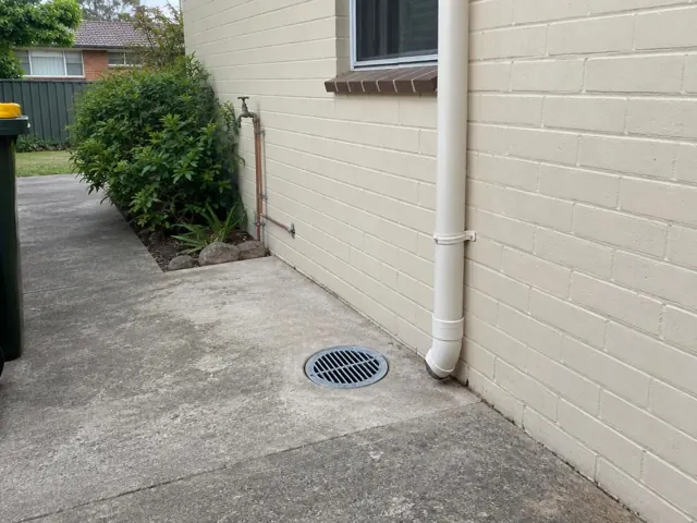 Outdoor drain grate set in a concrete path beside a cream brick house wall in Eastlakes
